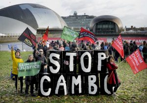 a group of people holding flags around a stack of black barrels painted with white letter spelling out stop cambo. the river clyde and the Glasgow science museum are visible in the background