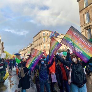 three people hold up rainbow flags reading no pride on a dead planet. they stand in front of a large march and there is a rain in the sky overhead