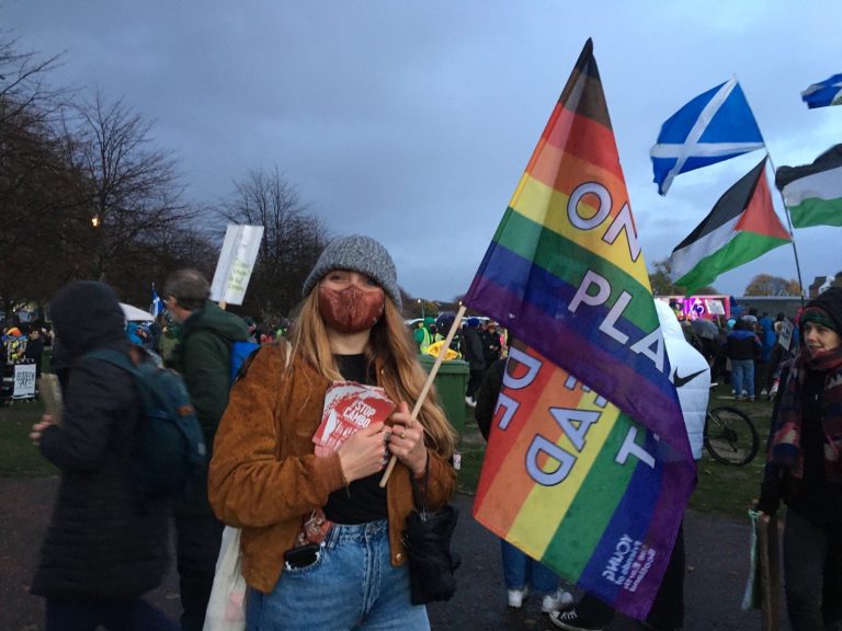 a young white woman in a mask holding a pride flag and stickers reading stop cambo. she is smiling stood in a crowd of people.