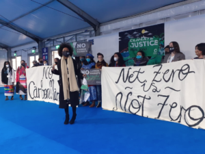 a row of people holding banners reading no carbon unicorns net zero is not zero climate justice now. A Black woman stands in front talking animatedly.