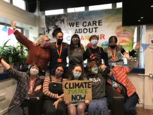 a group of happy looking young people wearing masks sit and stand around a sofa, holding a sign that says Climate action now 
