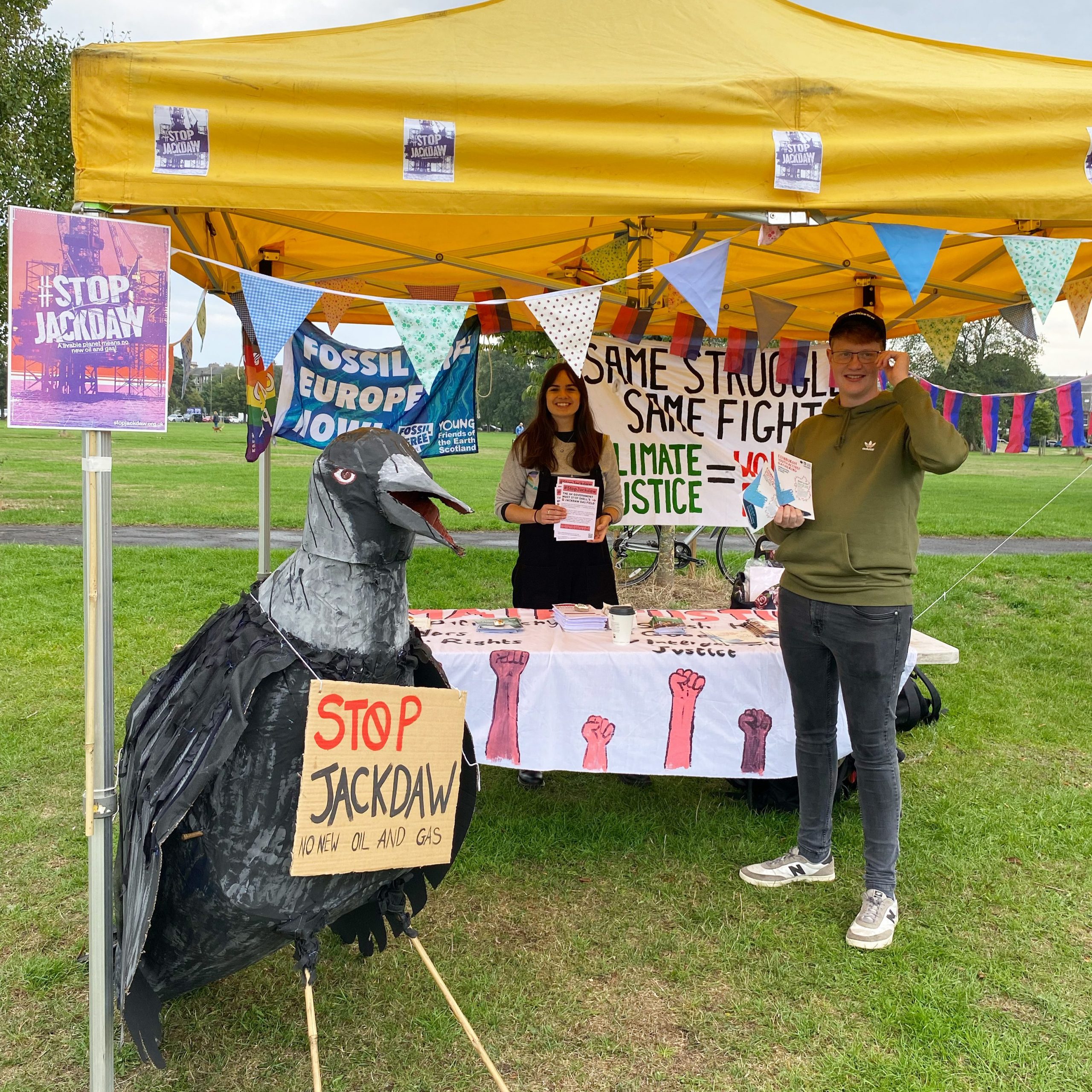two people and a giant paper mache model of a jackdaw under a yellow gazebo with a table full of flyers and banners