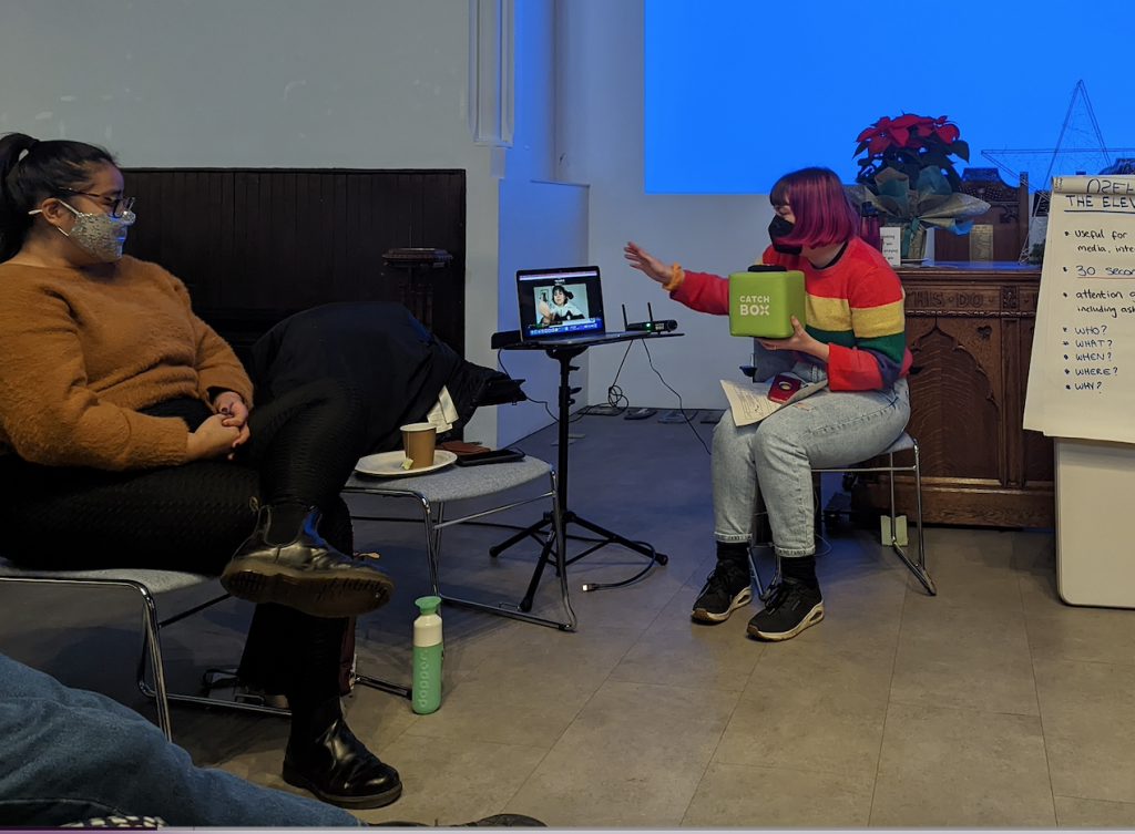 A person with pink hair wearing a mask and holding a green foam cube microphone gestures towards a laptop where another person is visible on Zoom. A third person wearing a mask watches on.