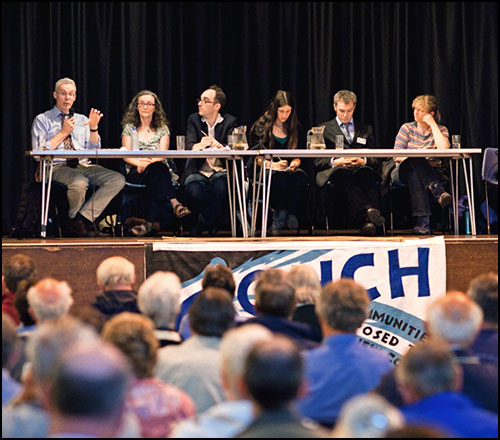 A group of people on a panel in front of a busy hall