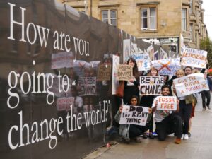 Students hold placards beside university hoarding reading 'How are you going to change the world today?'