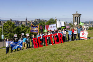 group of people with organisational banner and large letters spelling BETTER, located on calton hill overlooking Edinburgh