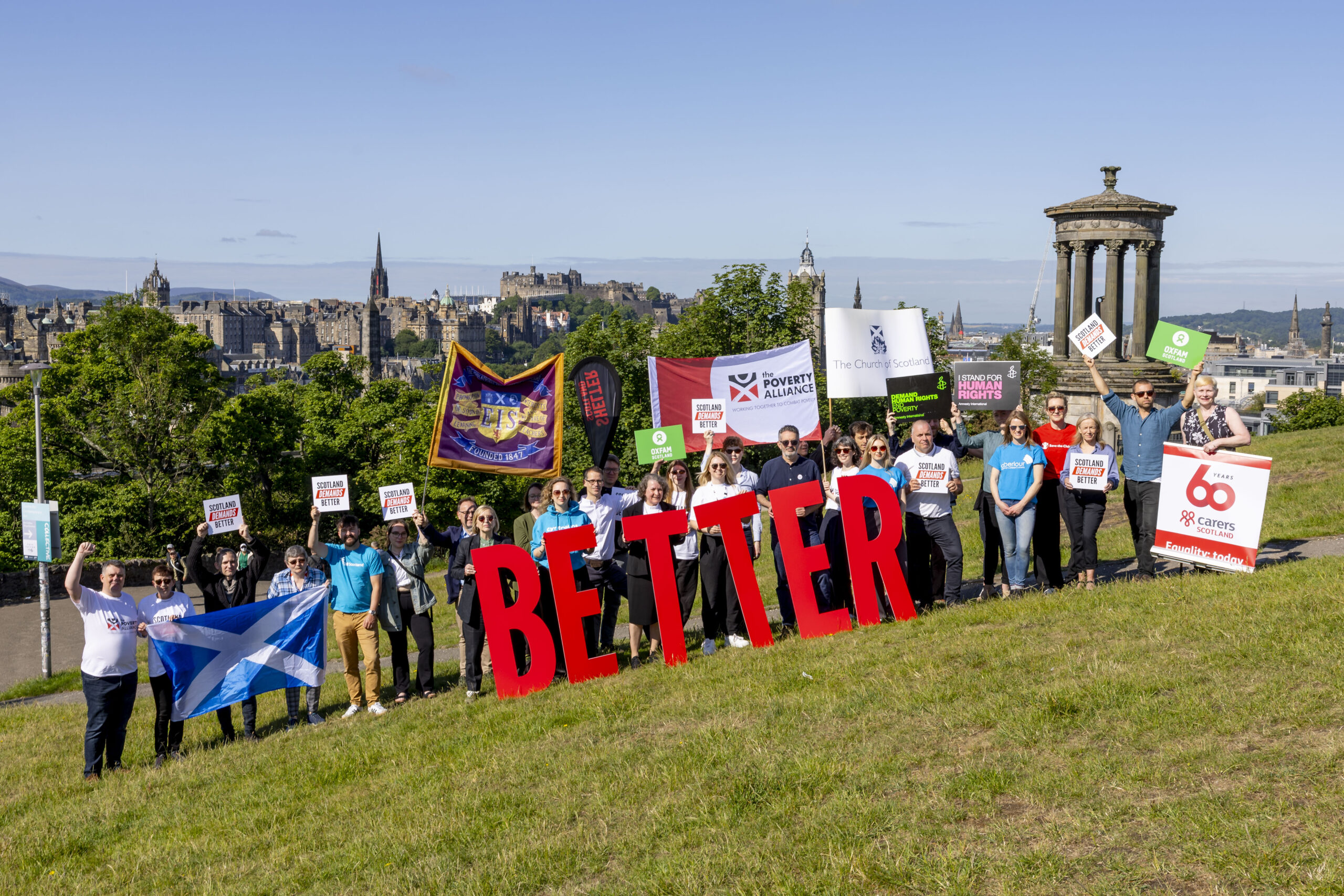 group of people with organisational banner and large letters spelling BETTER, located on calton hill overlooking Edinburgh