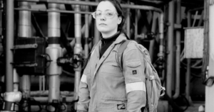 Black and white photo of a person looking directly at the camera, with industrial pipes in the background. The person is wearing overalls and glasses.