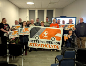 A group of people are holidng 'Better Buses for Strathclyde' campaign signs, in a room with a tv screen behind them.