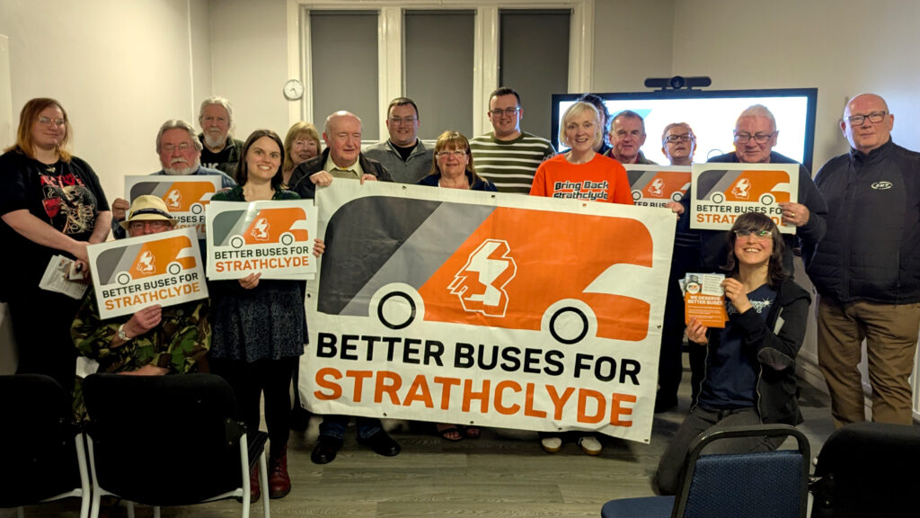 A group of people are holidng 'Better Buses for Strathclyde' campaign signs, in a room with a tv screen behind them.