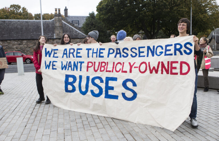 people standing behind a white banner saying "passengers want publicly owned buses"