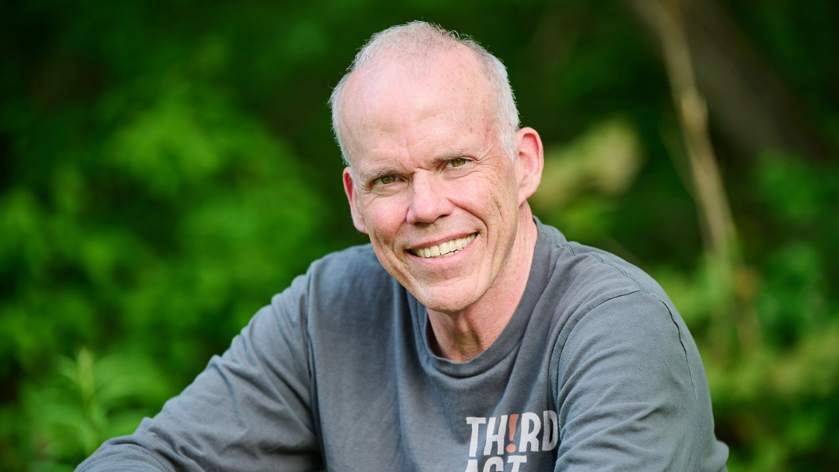 A man with a shaved head and a gray long-sleeved shirt smiles at the camera, with a blurred green background.