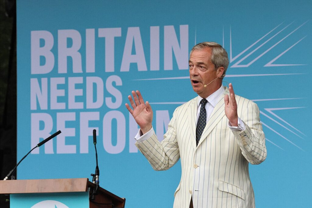 photo of nigel farage in white pin stripe suit speaking in front of a Reform banner